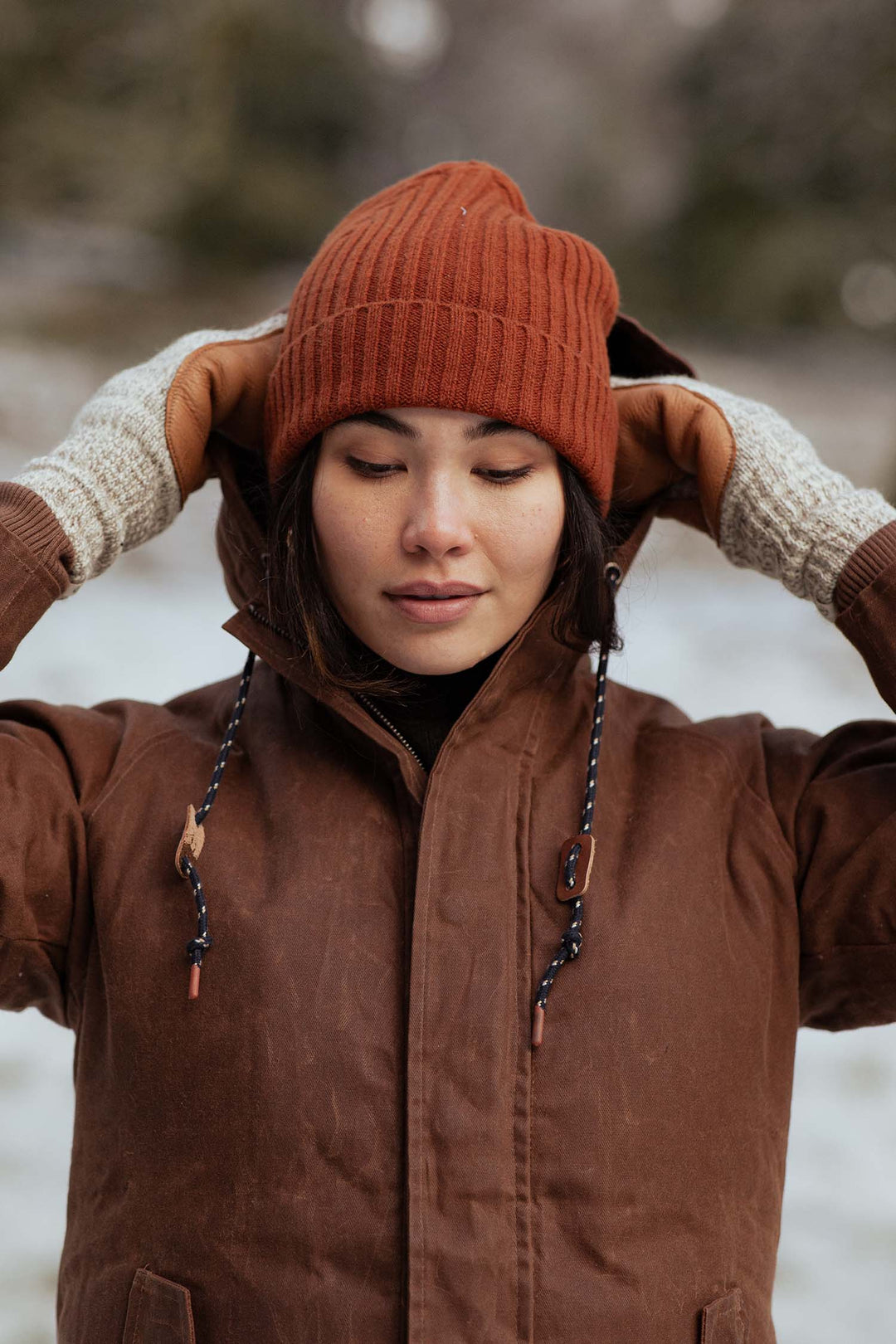 A person in a brown winter coat and beige gloves stands in the snow, gently holding the sides of their Bridge & Burn Chunky Ribbed Beanie in Sienna.