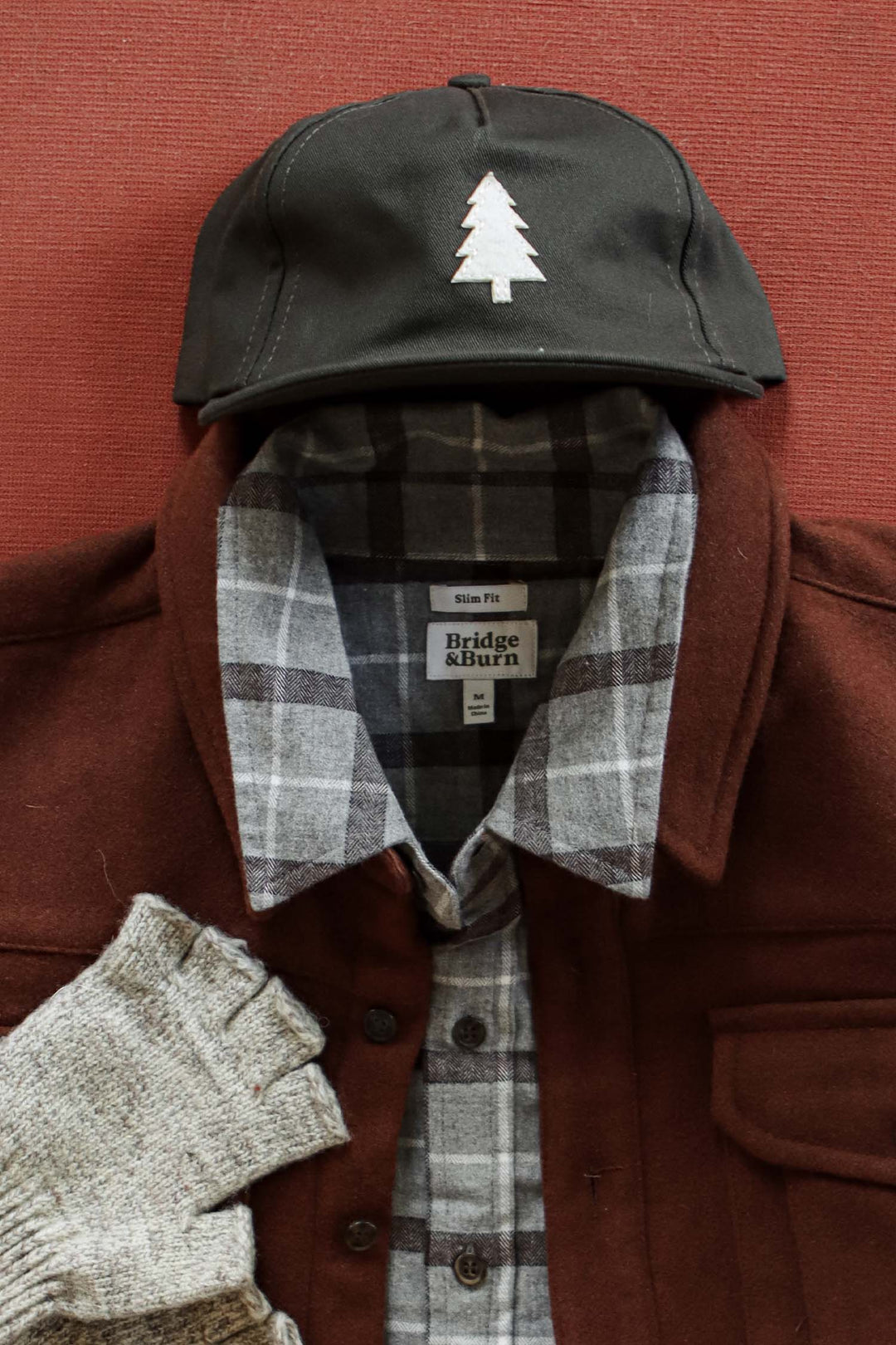 A flat lay on a reddish surface features a brown jacket over a gray plaid shirt, paired with Bridge & Burn’s Tree Cap in Olivewood, distinguished by its custom tree appliqué. Gray fingerless gloves are placed beside the jacket.