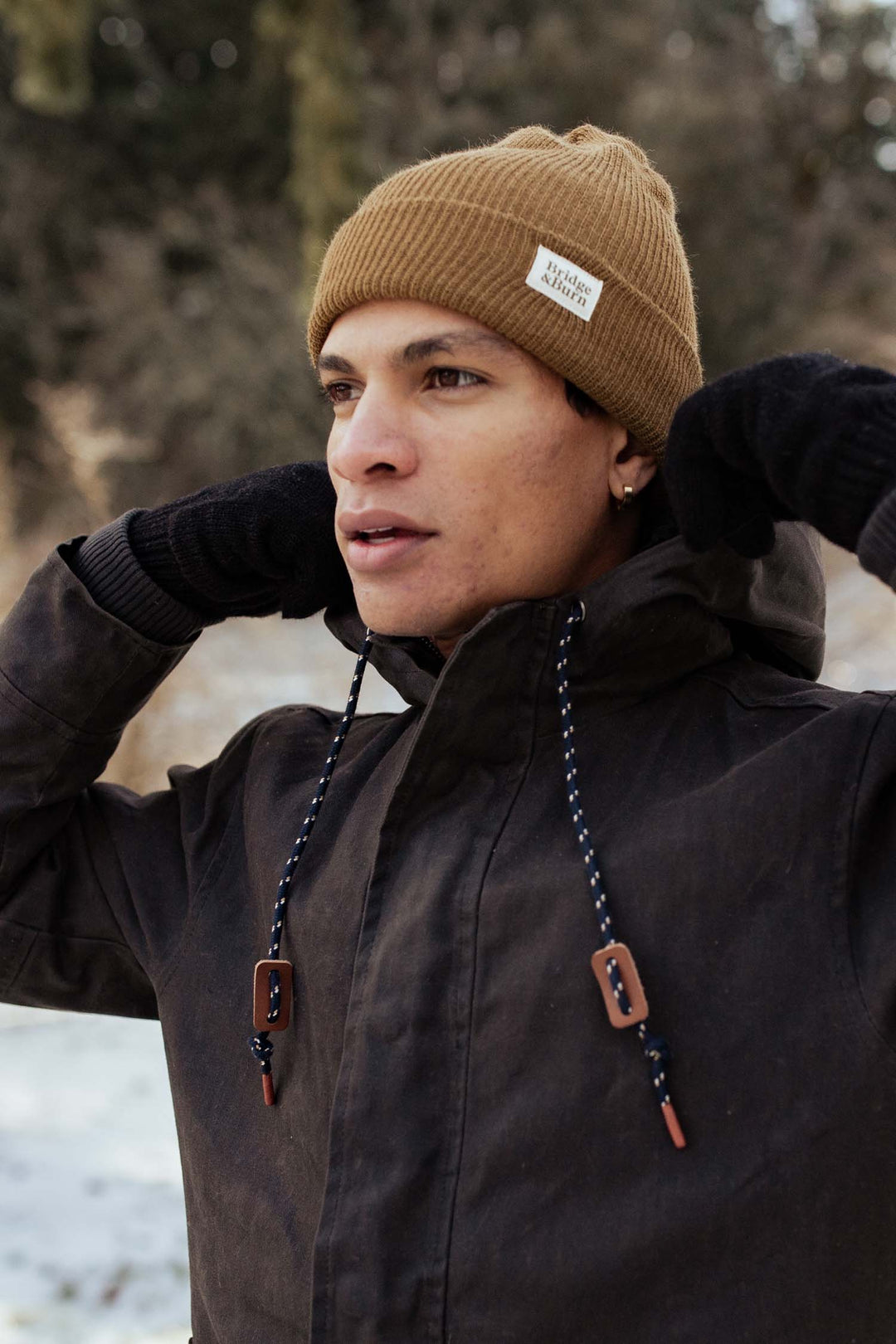 Wearing the Bridge & Burn Wool Watch Cap in Coyote, along with black gloves and a dark winter jacket, someone stands in a snowy, wooded area adjusting their hat and gazing into the distance.