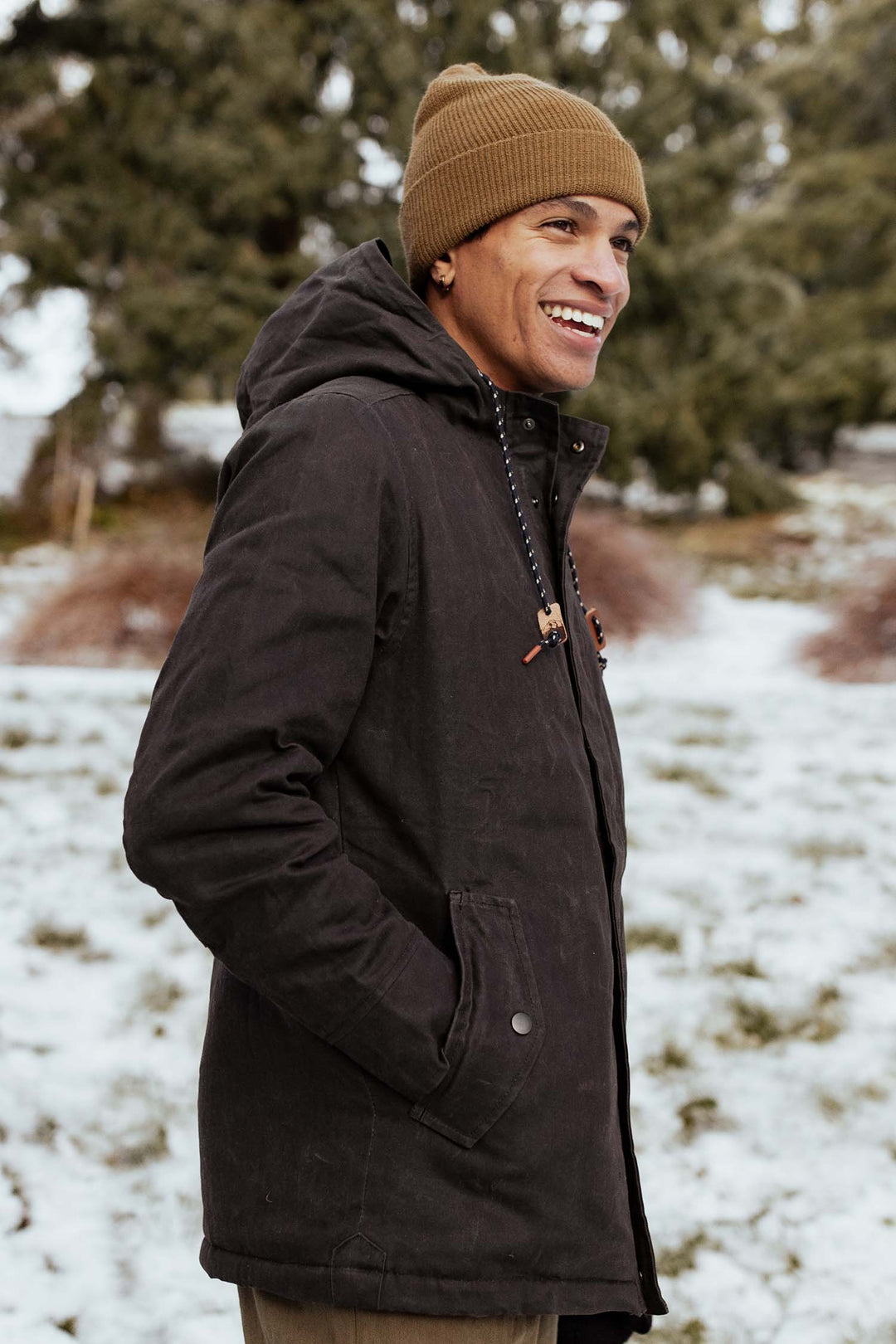 A person wears the Bridge & Burn Wool Watch Cap in Coyote and a dark winter jacket, standing outdoors among snowy evergreen trees, smiling and gazing to the side.