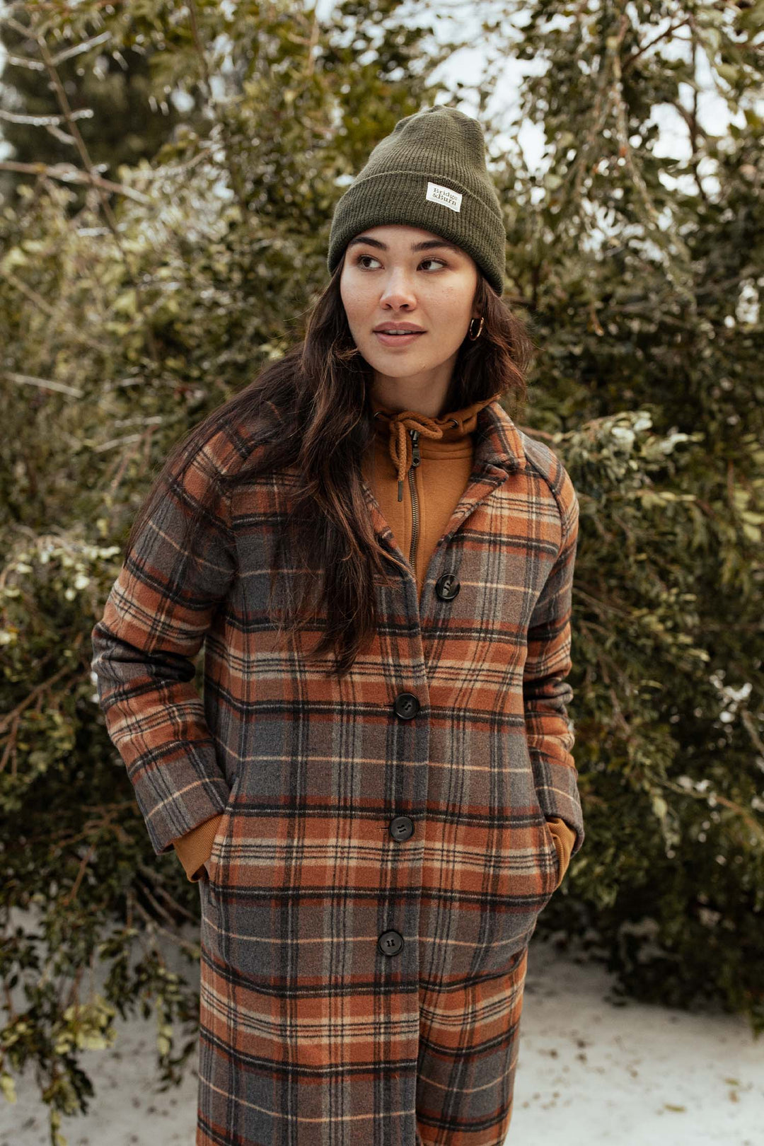 Wearing a Bridge & Burn Wool Watch Cap in Olive Drab, a person stands outside before snow-dusted greenery, hands tucked in their long plaid coat’s pockets, brown hoodie underneath, gazing thoughtfully to the side.