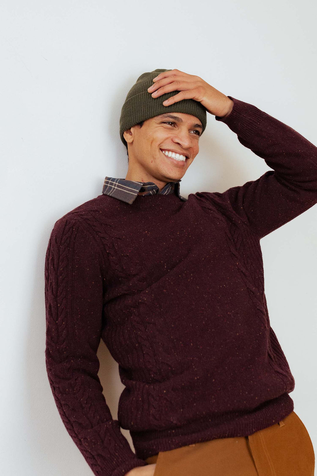 A smiling person in a dark maroon sweater, plaid-collared shirt, brown pants, and a Bridge & Burn Wool Watch Cap in Olive Drab made in the USA stands against a light wall, one hand touching their hat.