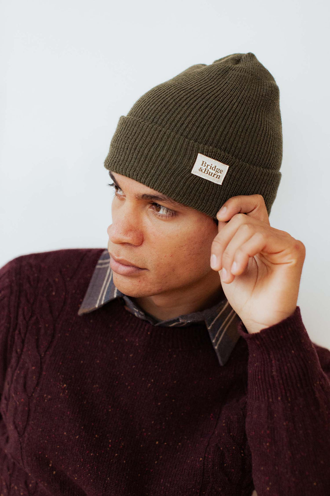 A person in a Bridge & Burn Olive Drab Wool Watch Cap and a maroon cable-knit sweater looks to the side while adjusting the beanie.