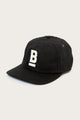 The B Flat Waxed Cap in black by Bridge & Burn features a white embroidered underlined letter B on the front, set against a plain light background.