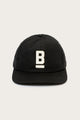 The B Flat Waxed Cap in black by Bridge & Burn features a white embroidered capital B with an underline on the front, set against a plain light background.