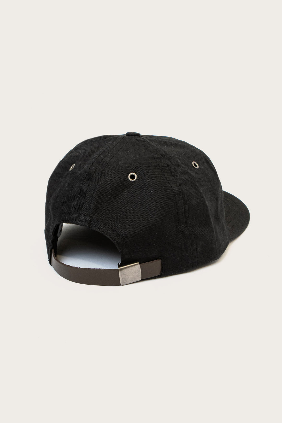 The Bridge & Burn B Flat Waxed Cap in Black features a metal buckle and adjustable strap at the back, metal eyelets for ventilation, and a curved brim—shown here against a simple light background.