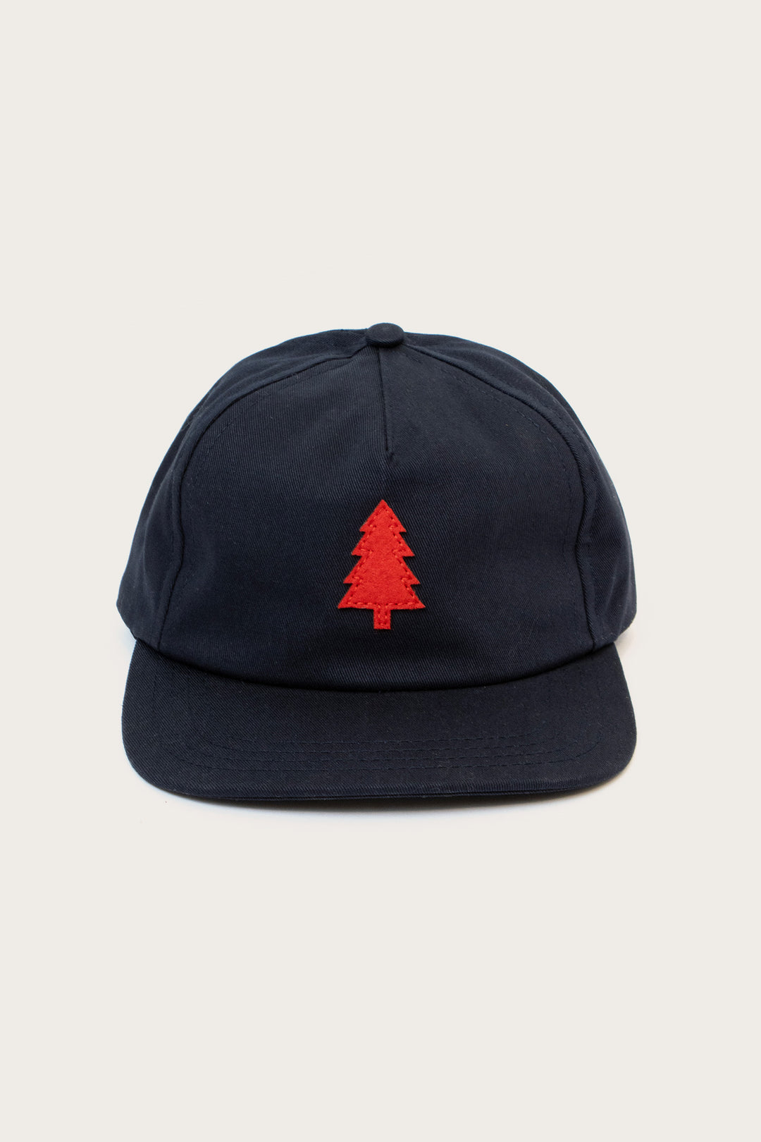 The Bridge & Burn Tree Cap in navy is a cotton twill hat featuring a red embroidered pine tree on the front and a leather size-adjustment strap, shown against a plain light background.