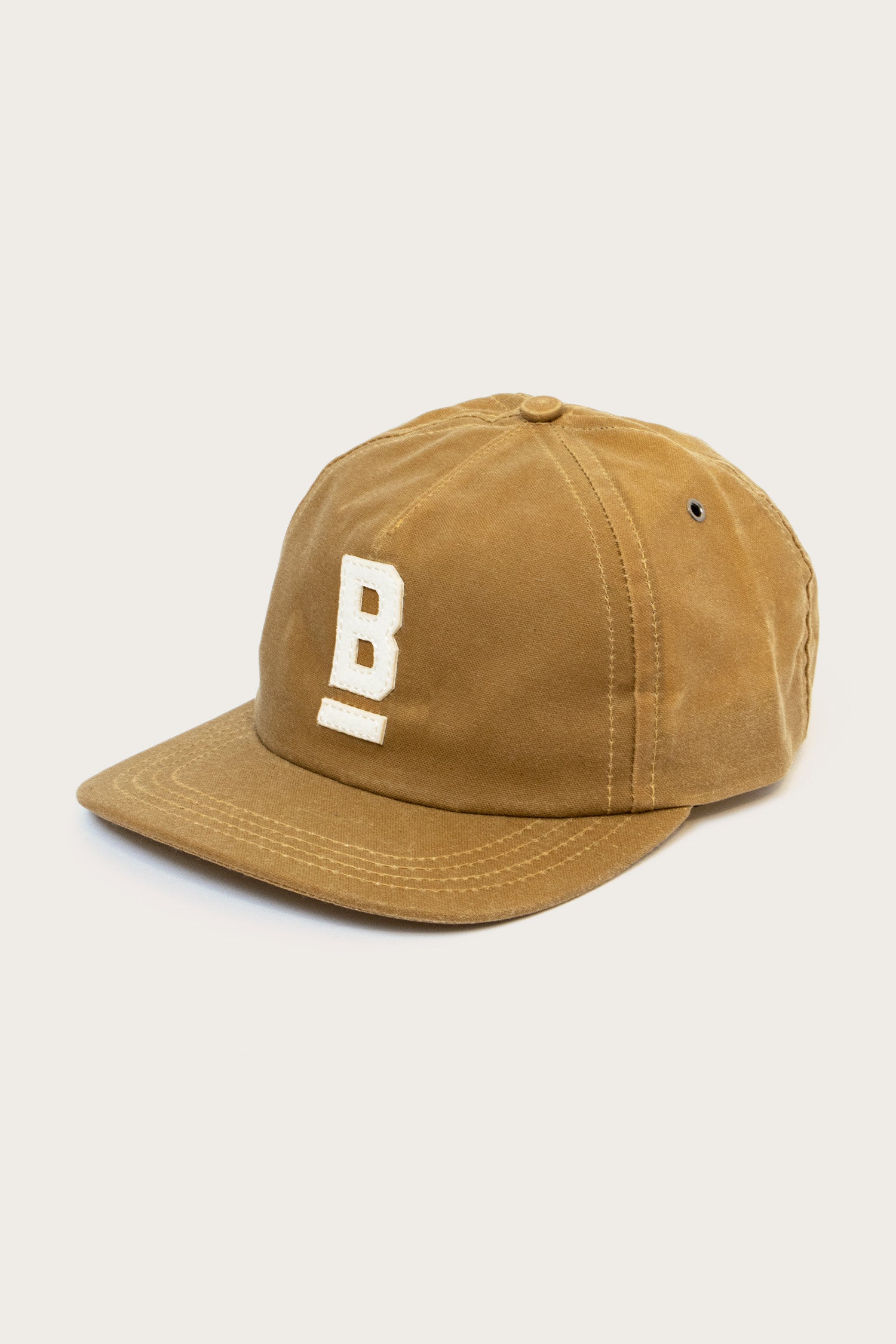 The Bridge & Burn B Flat Waxed Cap in brown is made from British Millerain waxed canvas, features a curved brim and bold white "B" embroidery on the front—perfect for wet weather.