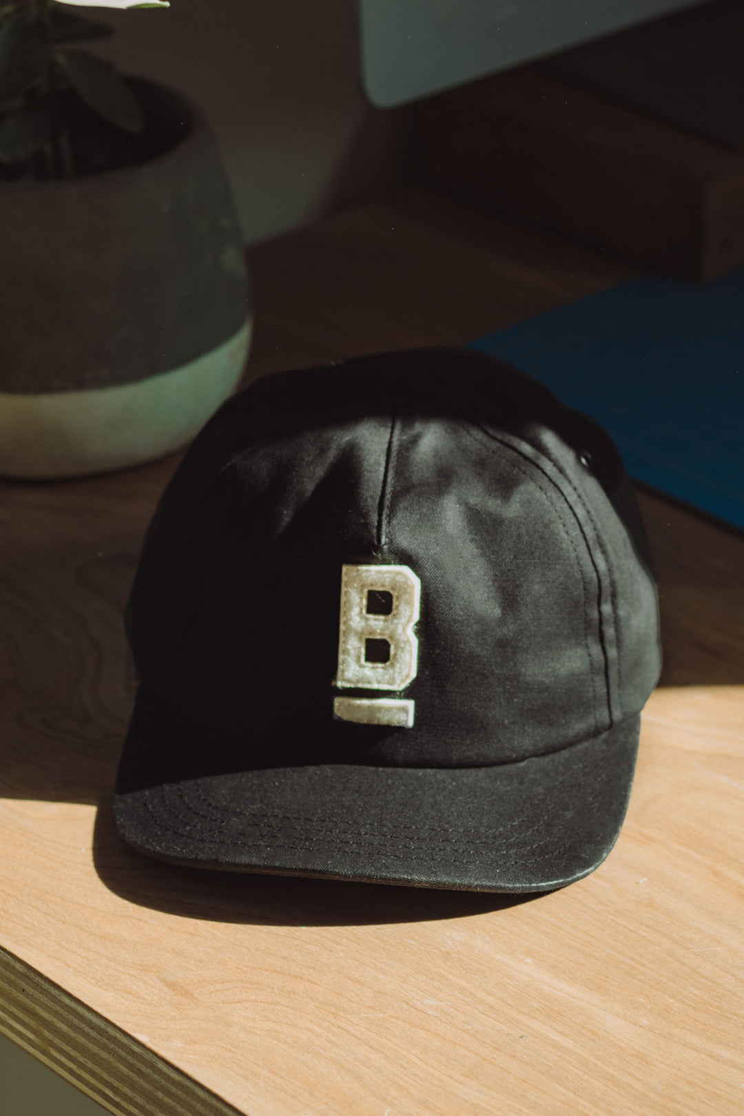 The Bridge & Burn B Flat Waxed Cap in Black, crafted from British Millerain waxed canvas and featuring a bold white B, sits on a wooden surface in sunlight with a blurred potted plant in the background.