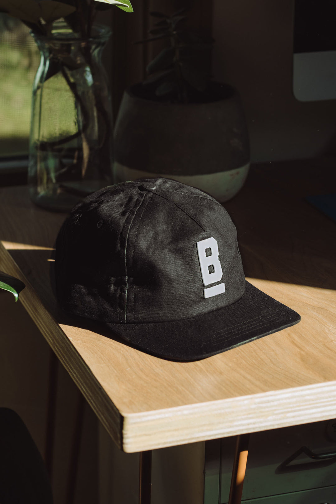 The Bridge & Burn B Flat Waxed Cap in black, made from British Millerain waxed canvas, showcases a bold white “B.” It’s styled on a sunlit wooden desk beside a glass jar and potted plant.