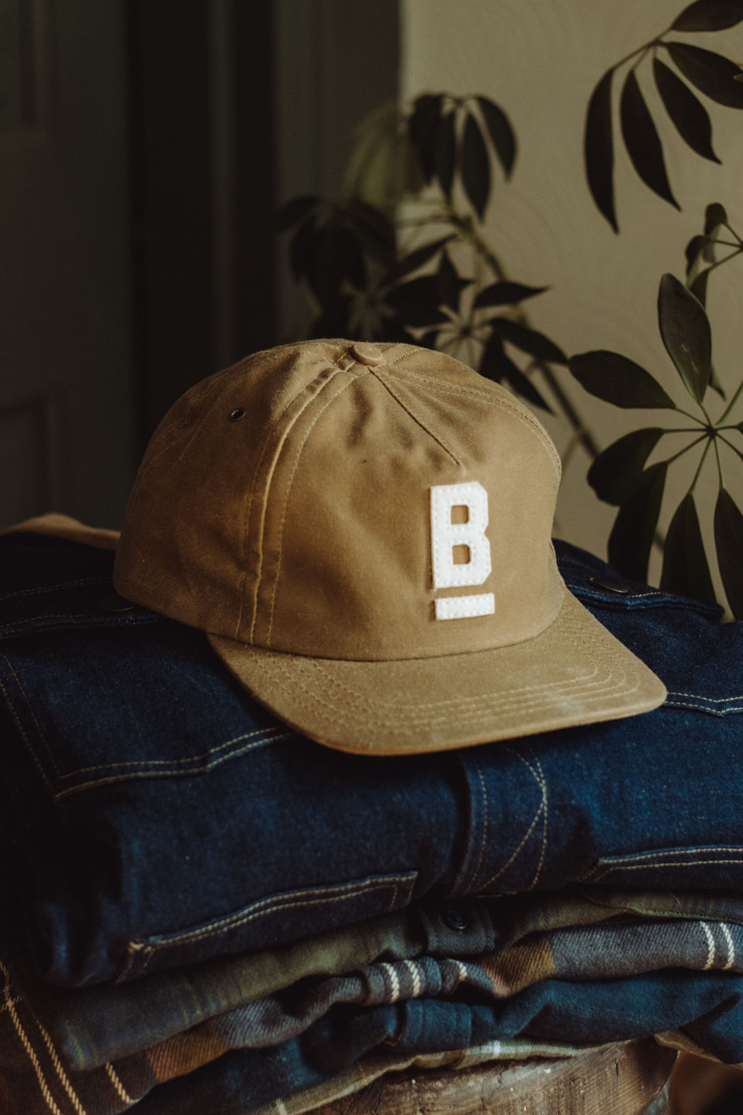 The Bridge & Burn B Flat Waxed Cap in brown, crafted from British Millerain waxed canvas, features a white "B" on the front. Shown atop folded blue denim with green plants behind—ideal for wet weather style.