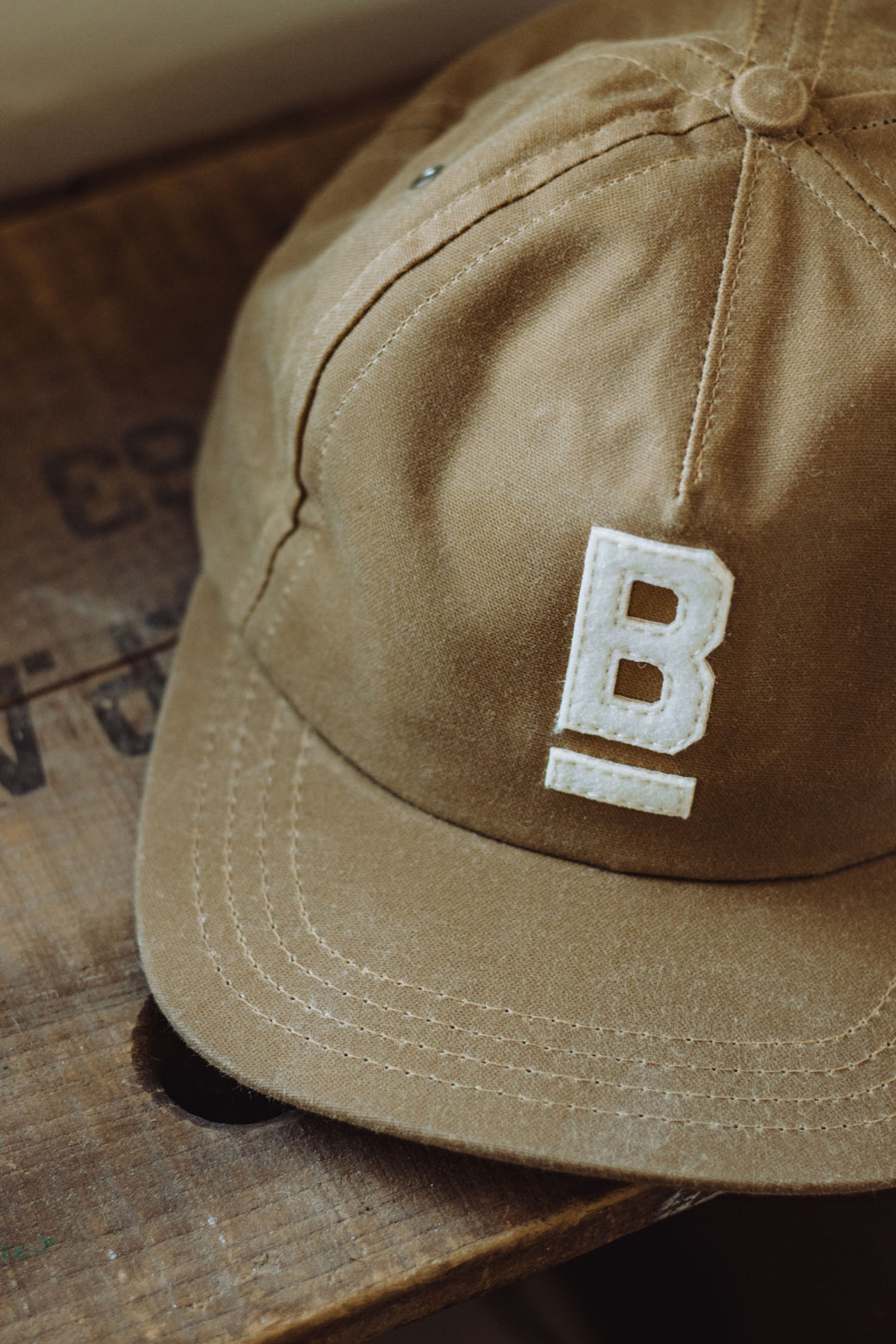 The B Flat Waxed Cap in brown by Bridge & Burn features British Millerain canvas and a white embroidered “B” with an underline on the front, displayed on a wooden surface.