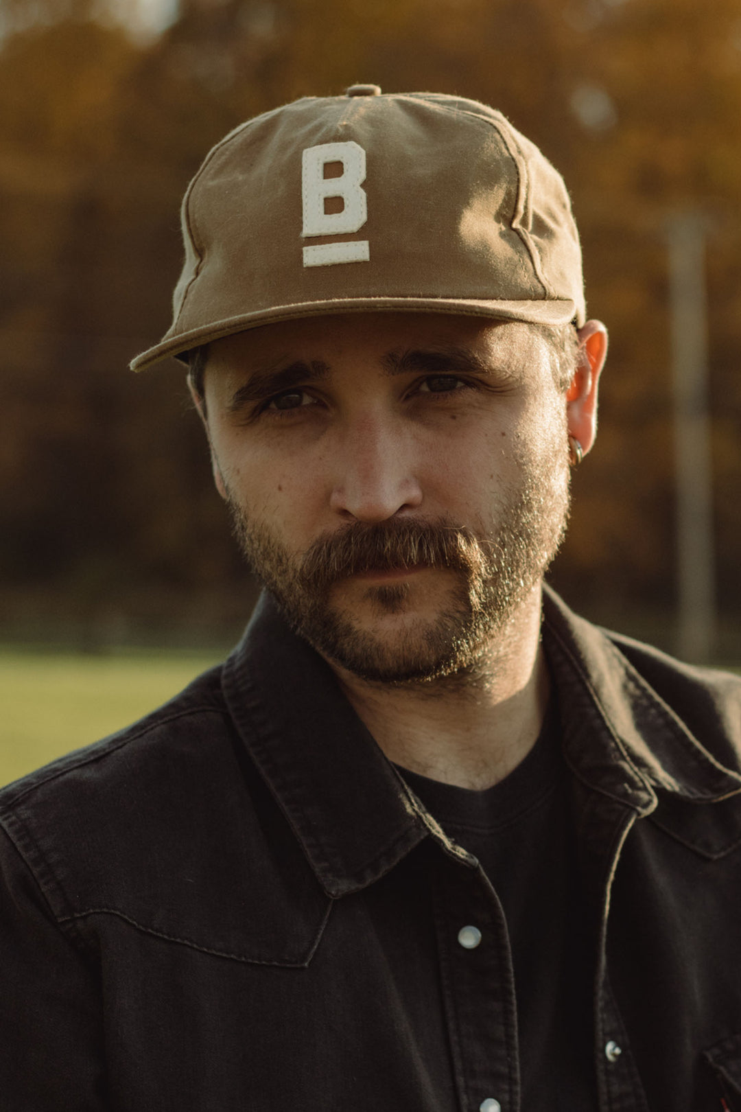 A man with a mustache and beard wears the Bridge & Burn B Flat Waxed Cap in brown—a classic British Millerain canvas hat with a white "B"—and a black jacket, standing outdoors with blurred autumn trees in the background.