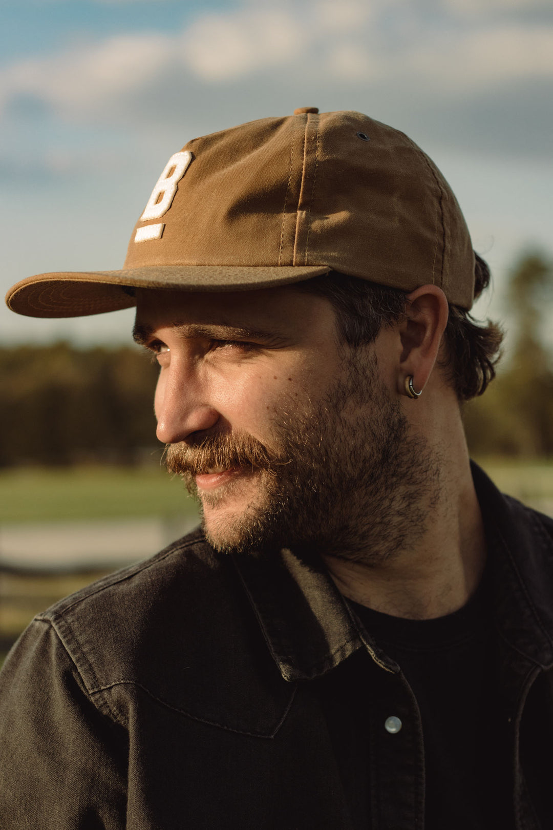 A man wears the Bridge & Burn B Flat Waxed Cap in brown, paired with a mustache, beard, dark jacket, and hoop earring. He looks to the side outdoors with trees and blue sky behind—a classic wet-weather style.