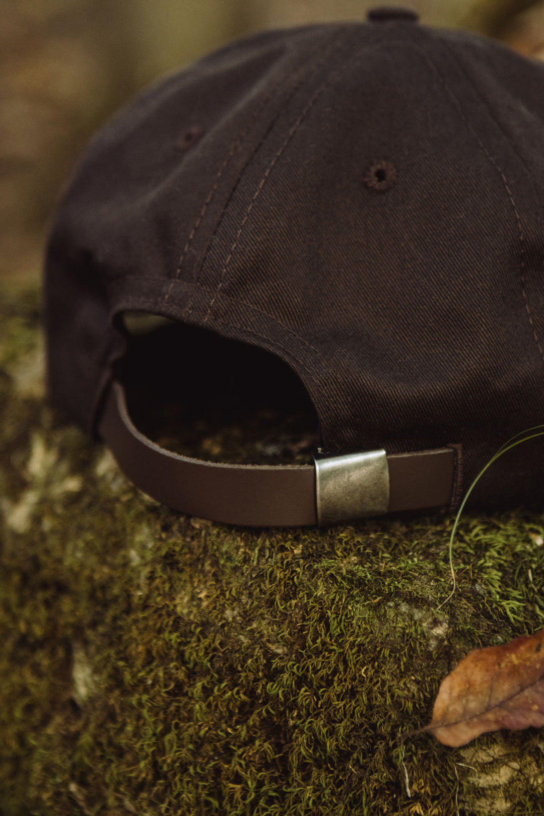 A close-up of the Bridge & Burn Tree Cap in Espresso, a brown cotton twill hat with an adjustable strap and metal clasp, resting on moss-covered rock outdoors.