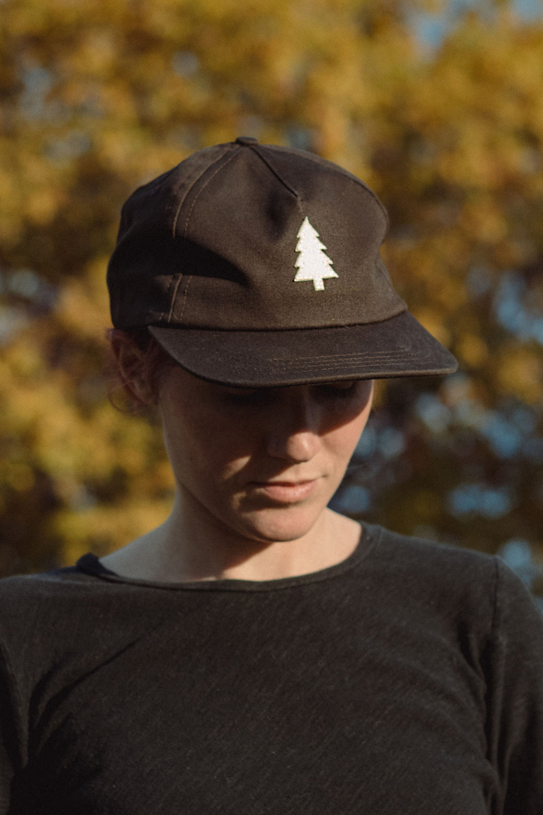 A person wearing the Bridge & Burn Tree Cap in Espresso, featuring a white tree emblem, looks down while standing outdoors with blurred autumn trees in the background.