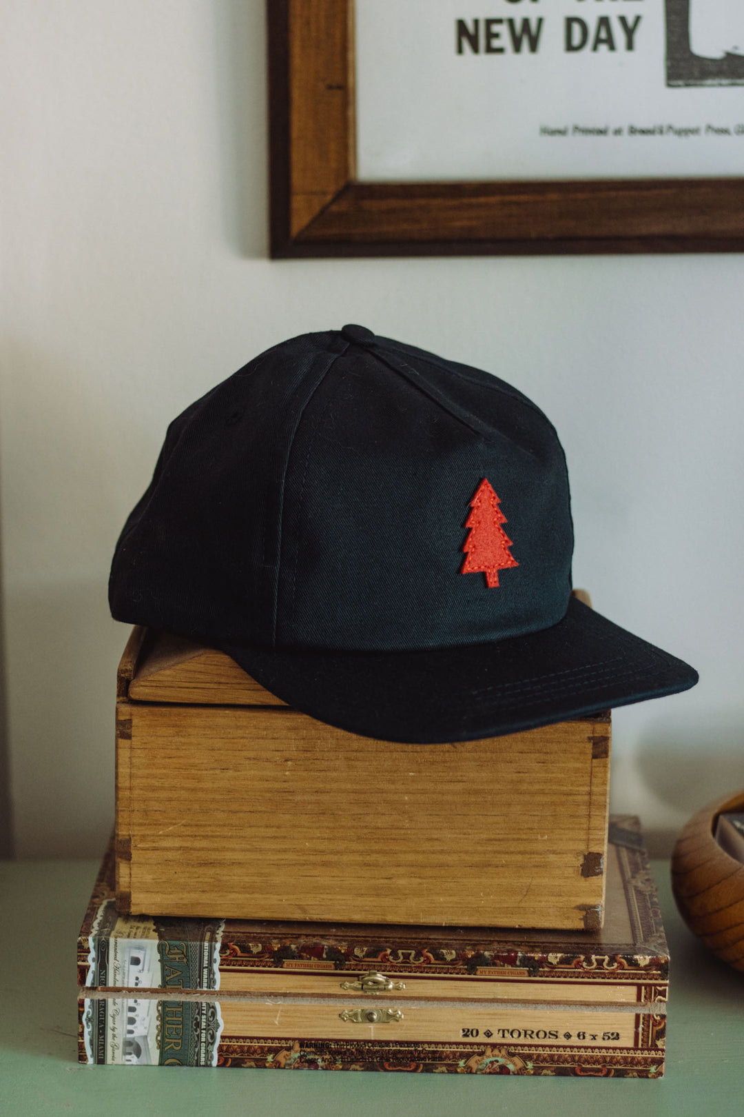 A navy Tree Cap by Bridge & Burn, featuring a red tree emblem and a leather adjustment strap, rests on a wooden box atop two books, with part of a framed picture visible in the background.