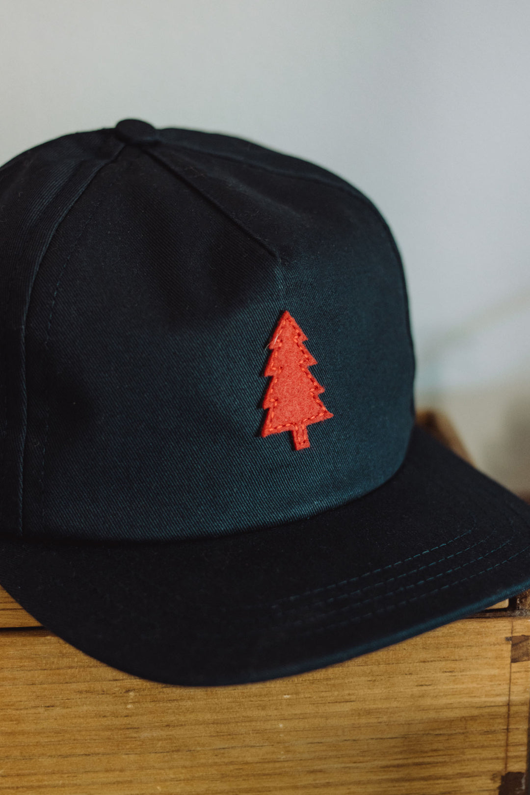 A navy cotton twill Tree Cap by Bridge & Burn, featuring a red embroidered pine tree emblem and a leather adjustment strap, is displayed on a wooden surface.
