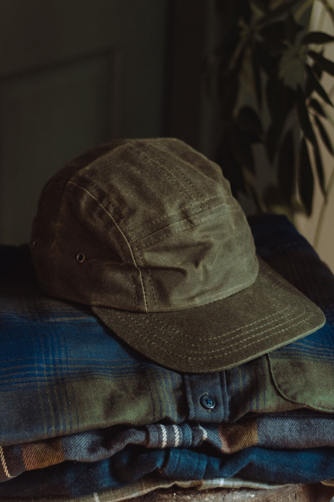 The Bridge & Burn Camper Waxed 5 Panel Cap in Moss, made in the USA, sits atop a stack of blue and brown plaid shirts, softly lit with a blurred leafy plant in the background.