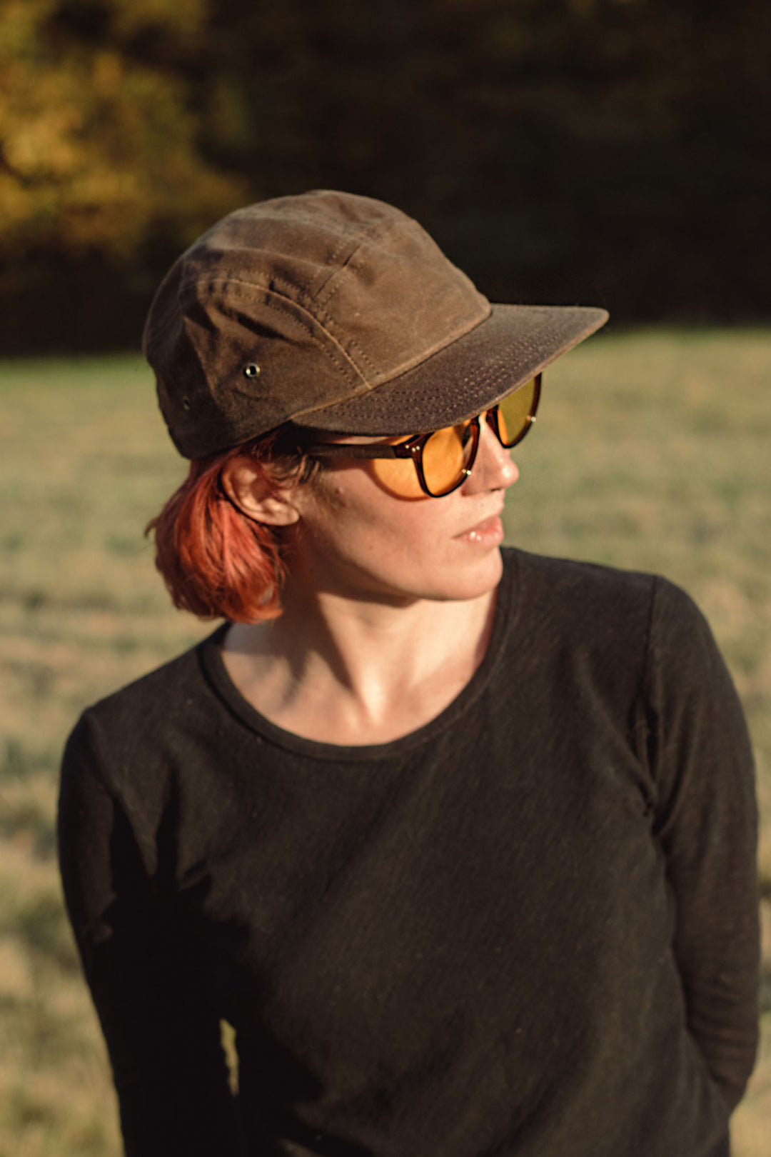 A person with short red hair wears the Bridge & Burn Camper Waxed 5 Panel Cap in Umber, amber sunglasses, and a black long-sleeve shirt while standing in a grassy field with blurred trees and sunlight in the background.