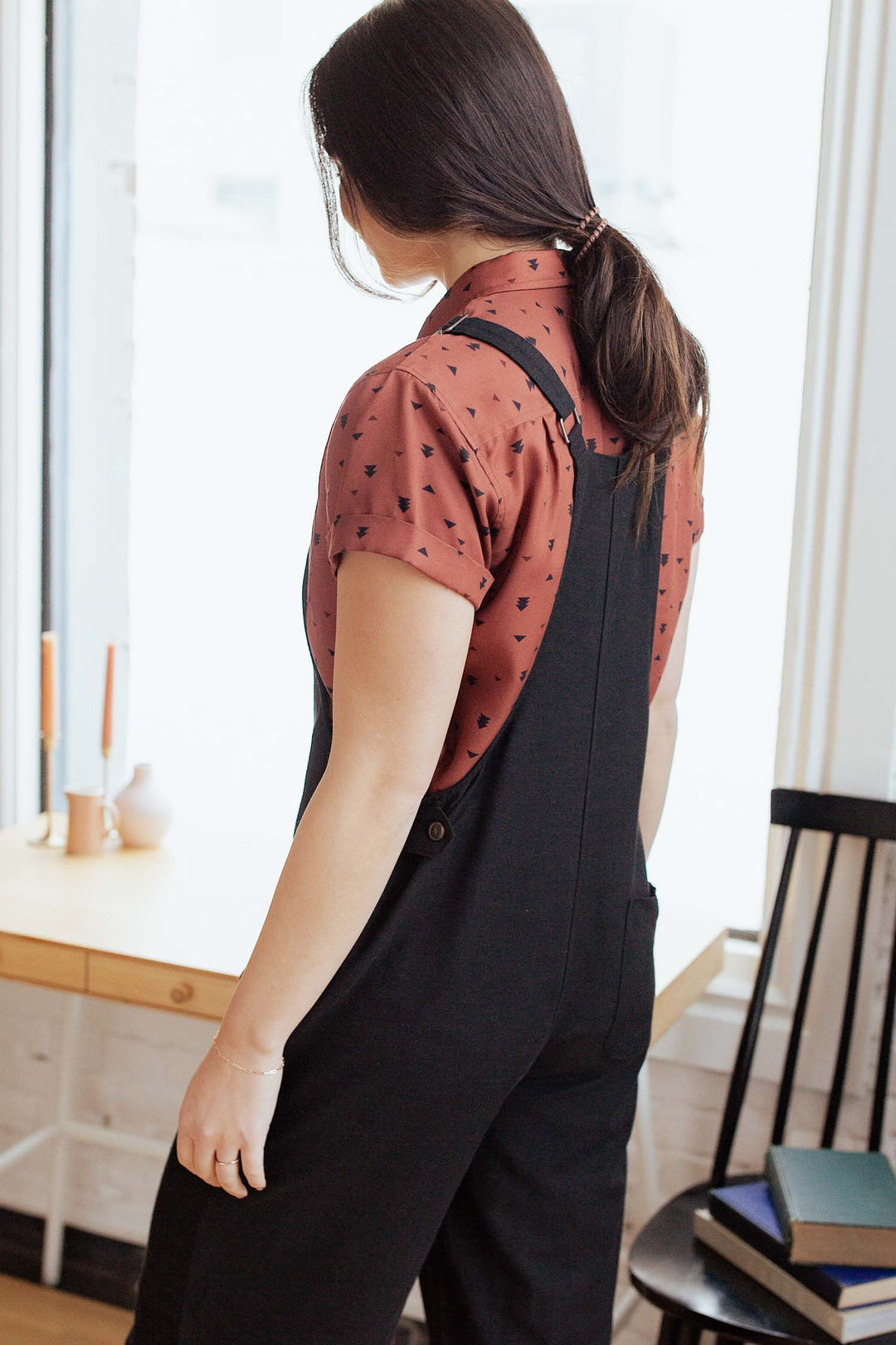 A person with long dark hair in a low ponytail stands indoors, wearing a short-sleeve, patterned rust shirt and the Bridge & Burn Edie Overall in Black, which features adjustable straps and wide legs, facing a window near a wooden desk adorned with candles and accompanied by a chair.