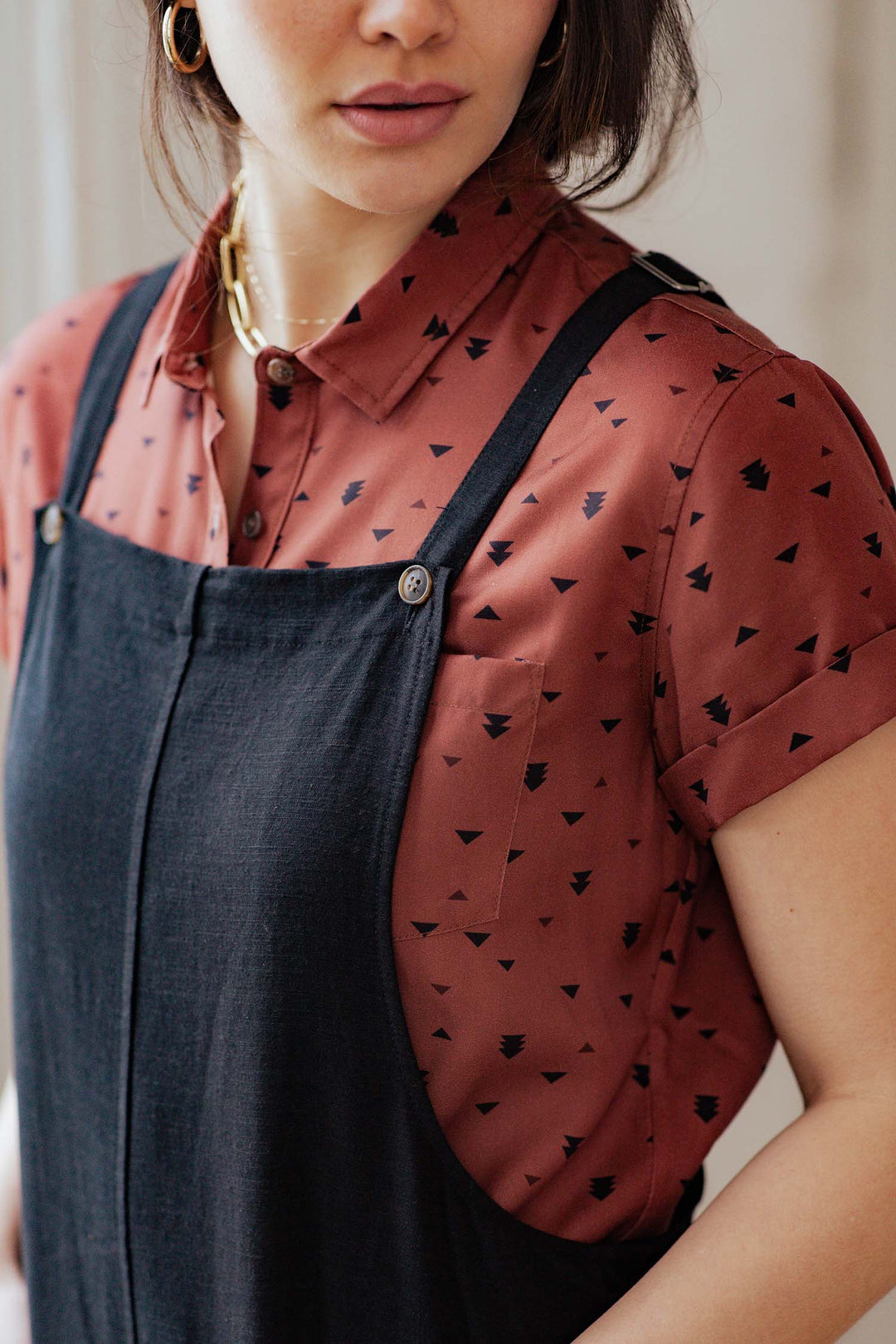 A woman wearing the Bridge & Burn Edie Overall in Black, featuring adjustable straps, layered over a rust-colored, short-sleeve button-up shirt with black triangle patterns. Only her lower face and upper torso are visible.
