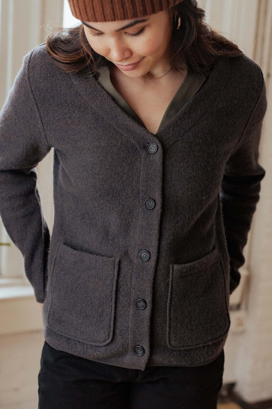 A woman wearing a brown beanie and the Bridge & Burn Whit Cardigan in Charcoal with patch pockets gazes down gently near a softly lit window indoors.