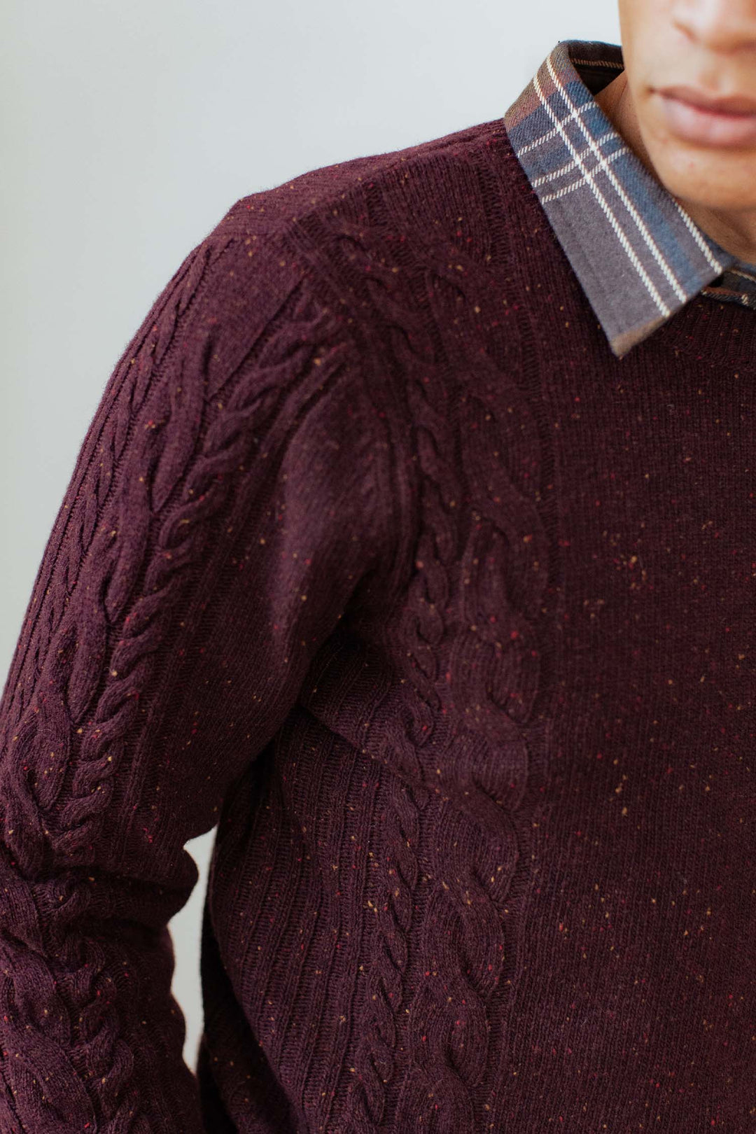 A person wears the Bridge & Burn Hugo Sweater in Burgundy Donegal—a maroon crewneck with cable detail and a flecked pattern—over a plaid collared shirt; only their lower face, shoulder, and upper torso are visible.