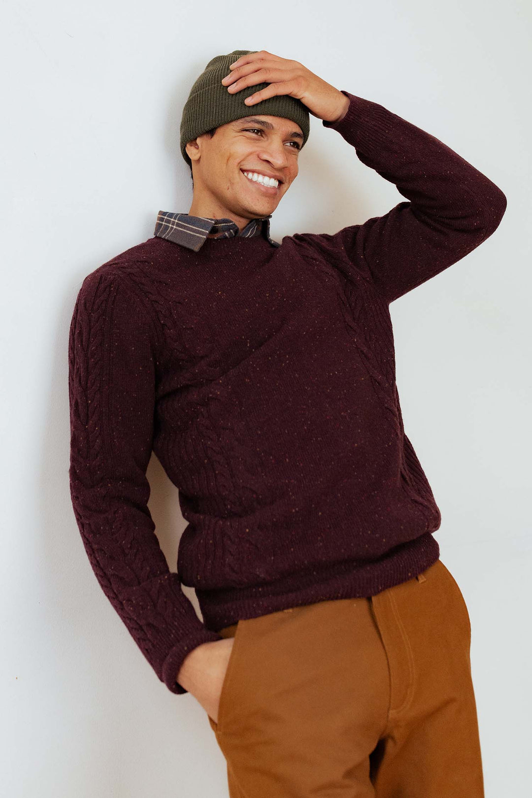 A smiling person wears the Bridge & Burn Hugo Sweater in Burgundy Donegal over a plaid shirt, paired with brown pants and a green beanie, standing against a white wall with one hand in their pocket and the other touching their head.