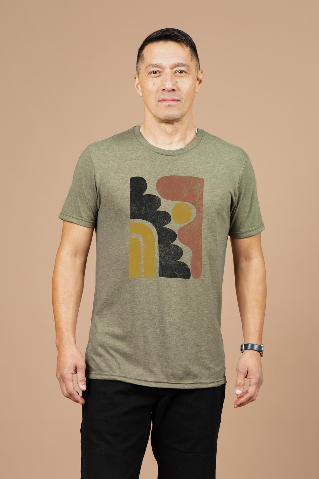 A man stands against a beige background, wearing Bridge & Burn’s Men's Ladera Tee in olive—a green organic cotton shirt featuring an abstract geometric design in black, brown, and gold—paired with black pants and a wristwatch.