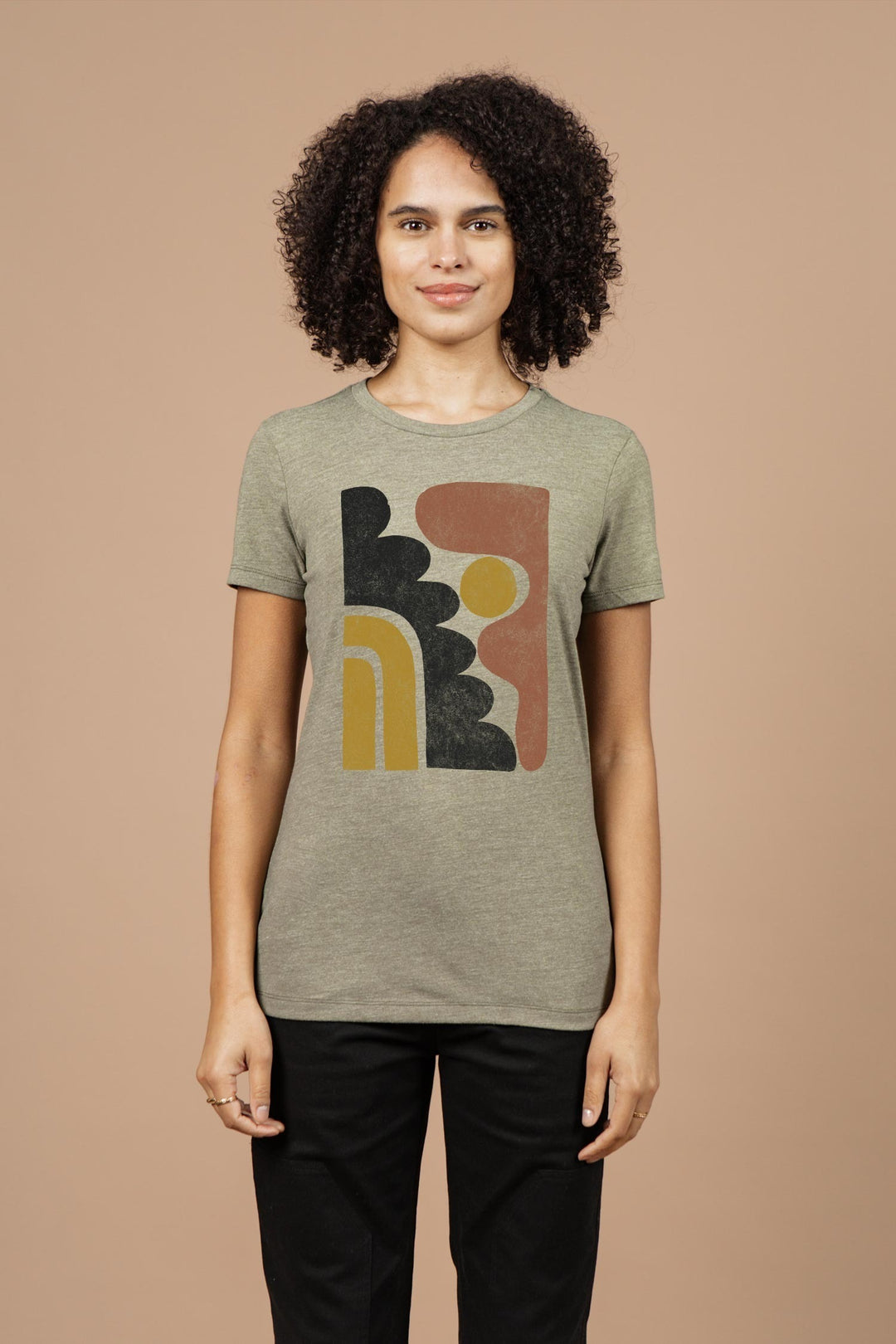 A person with curly hair wears the Bridge & Burn Women’s Ladera Tee in olive, an organic cotton lightweight top with a black, yellow, and rust geometric design, standing against a plain beige background.