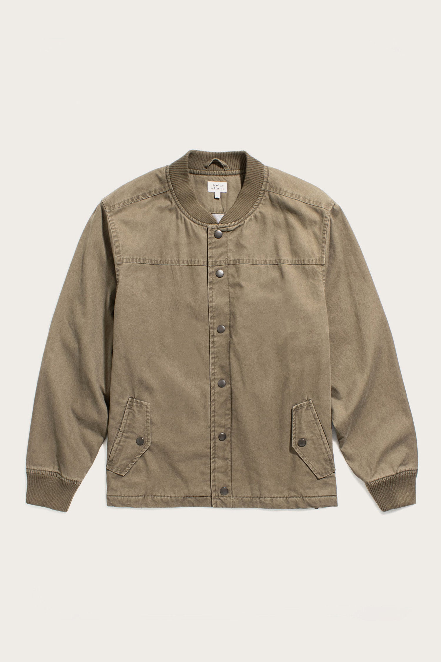 The Bridge & Burn Austen Varsity Jacket in khaki green canvas features long sleeves, snap buttons, ribbed cuffs and collar, and two front flap pockets, shown laid flat on a light background.