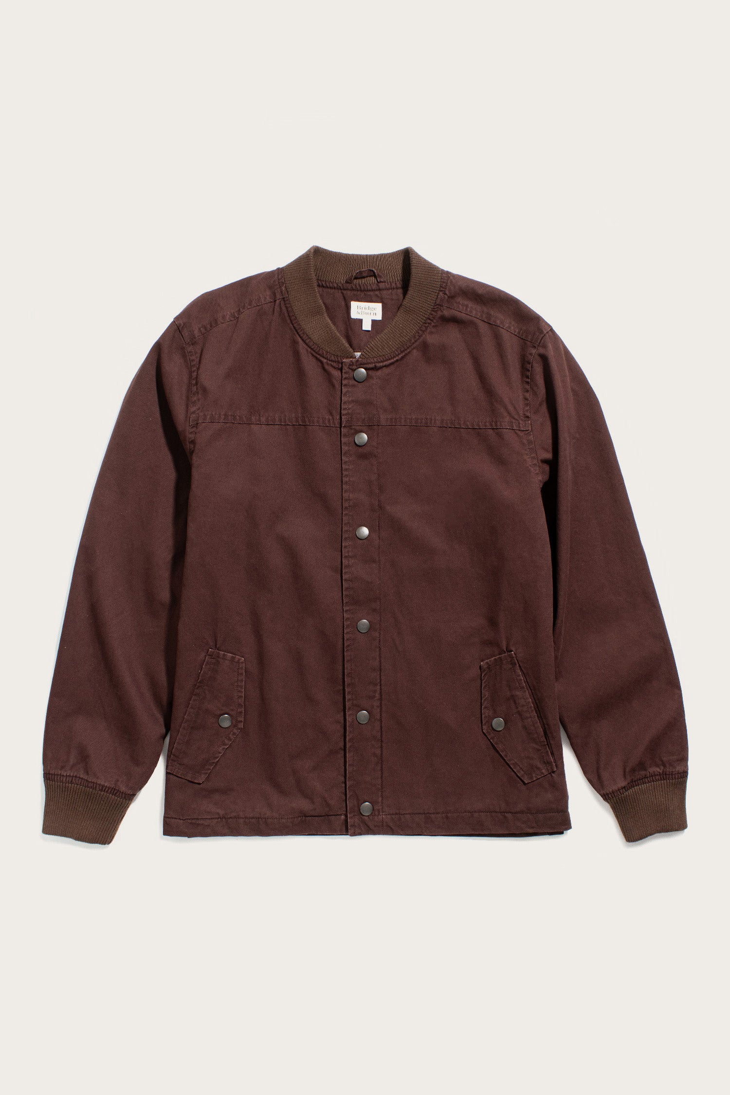 The Austen Varsity Jacket in mahogany canvas by Bridge & Burn features a cotton shell, long sleeves, ribbed trim, two button-flap front pockets, and a relaxed fit, displayed on an off-white background.