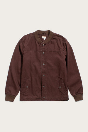 The Austen Varsity Jacket in mahogany canvas by Bridge & Burn features a cotton shell, long sleeves, ribbed trim, two button-flap front pockets, and a relaxed fit, displayed on an off-white background.