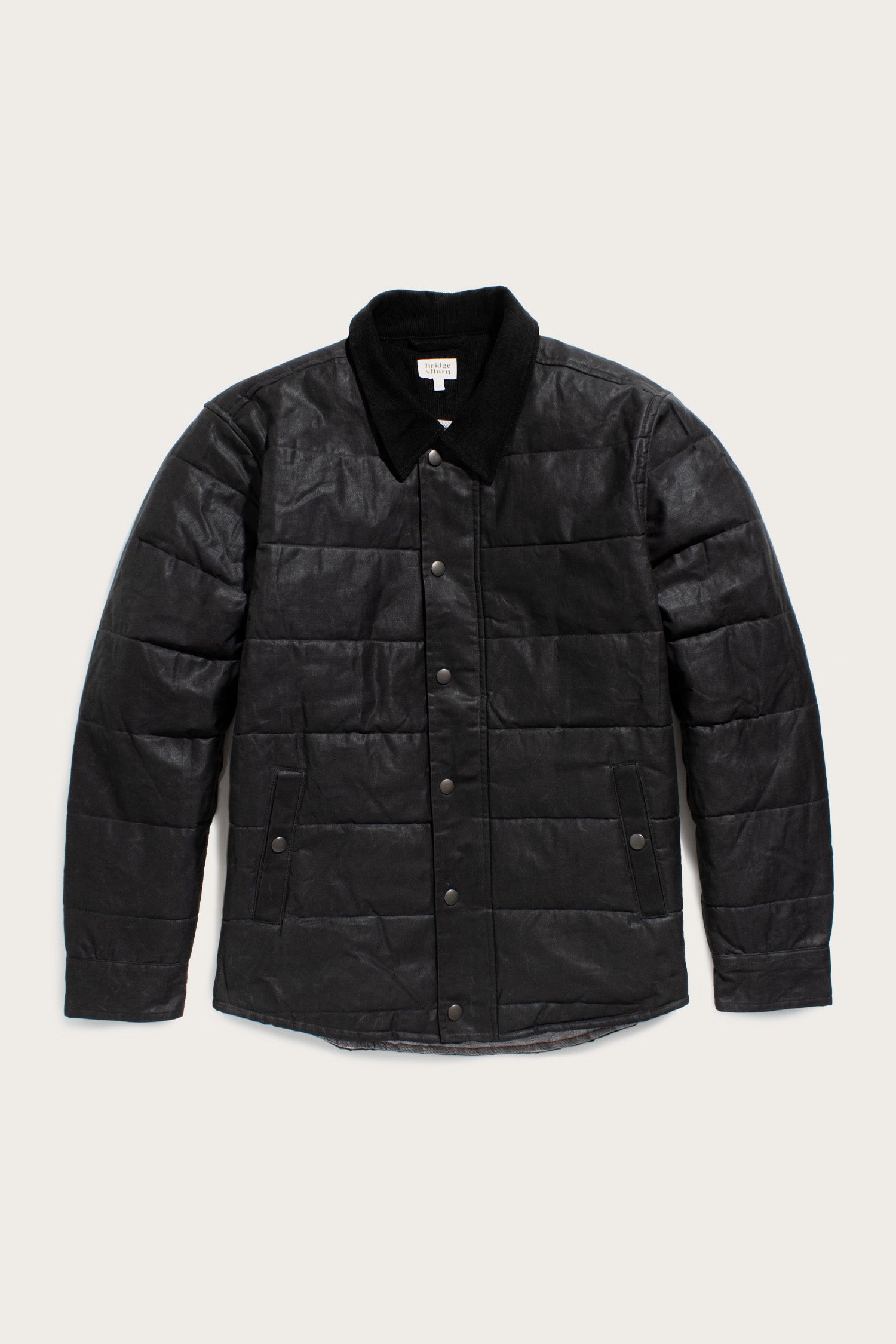 The Bridge & Burn Holman Quilted Jacket in black waxed canvas features a quilted design, button-up front, long sleeves, and a soft collar, shown on a light background.