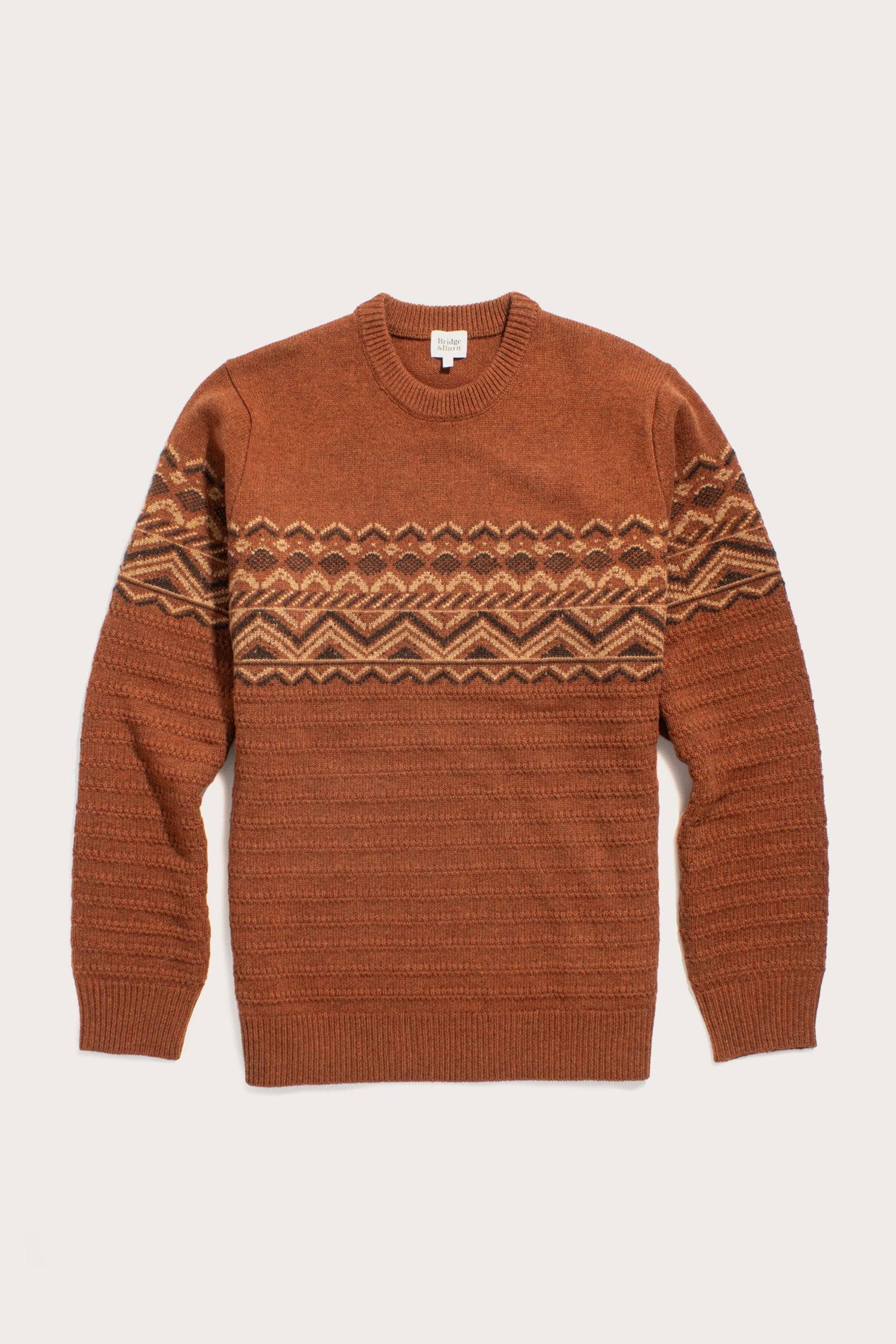 The Bridge & Burn Rockwood Fairisle Sweater in Walnut Grove is a rust-colored merino wool knit with a crew neck, long sleeves, and a beige and brown geometric fairisle pattern across the chest and upper arms.