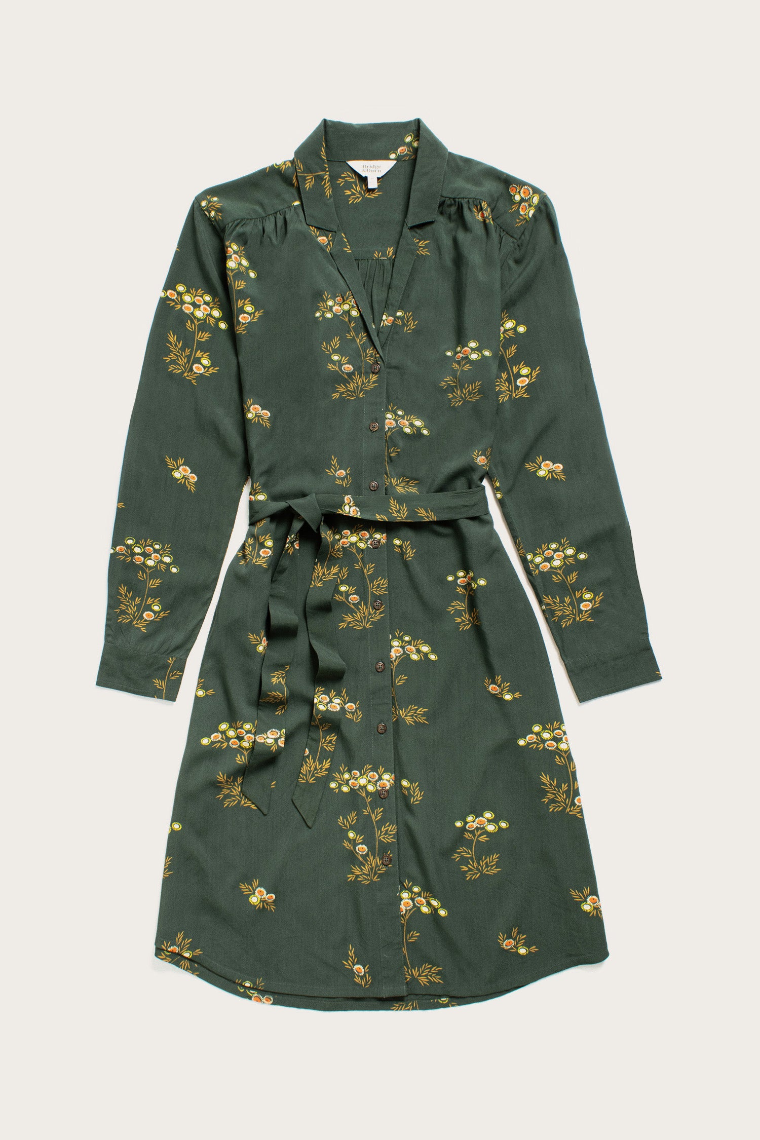 The Bridge & Burn Emery Shirt Dress in Woodland Bloom—a long-sleeve, knee-length shirtdress with a yellow and white floral print, button-down front, collar, and waist tie—is shown laid flat on a light background.