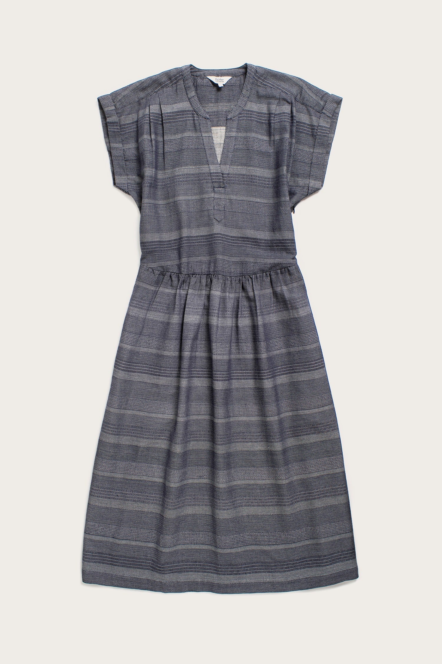 The Bridge & Burn Ira Dress in Cascade Weave is a short-sleeve, knee-length organic cotton dress with subtle gray stripes, V-neckline, gathered waist, and a relaxed pullover fit for effortless comfort.