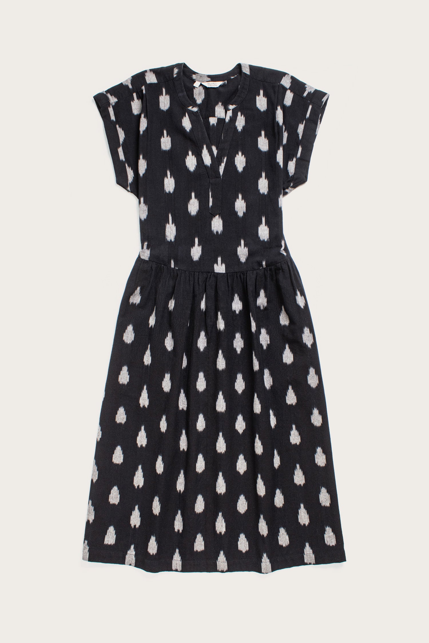 The Ira Dress in Eclipse Ikat by Bridge & Burn is a black, knee-length pullover with short dolman sleeves, V-neck, and gathered waist. It features an abstract white dotted pattern and is displayed flat on a white background.