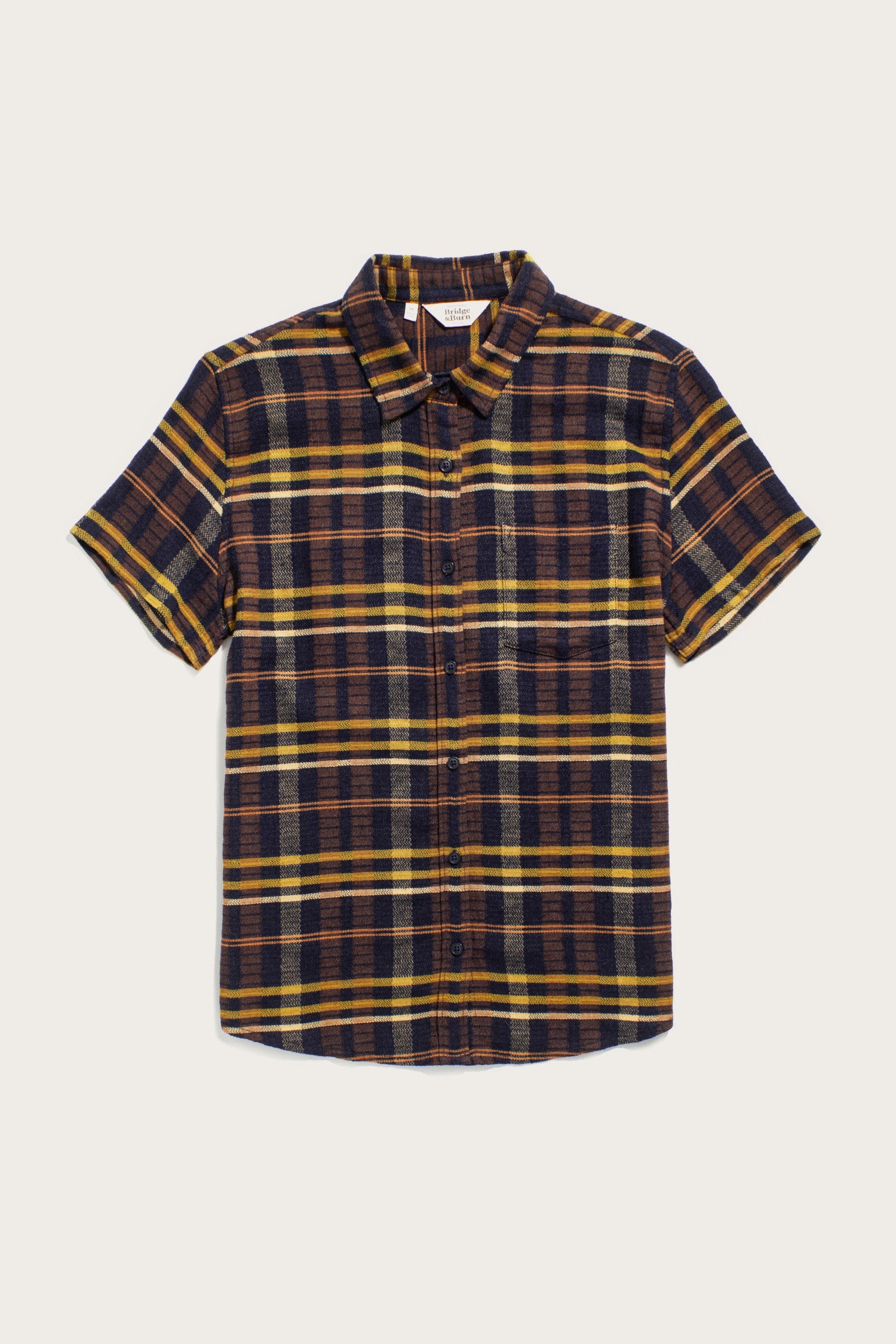 The Bridge & Burn Lana Shirt in Desert Dawn Plaid features a relaxed fit, short sleeves, collar, front chest pocket, and is crafted from organic cotton in yellow, brown, and navy blue plaid. Shown laid flat on a white background.