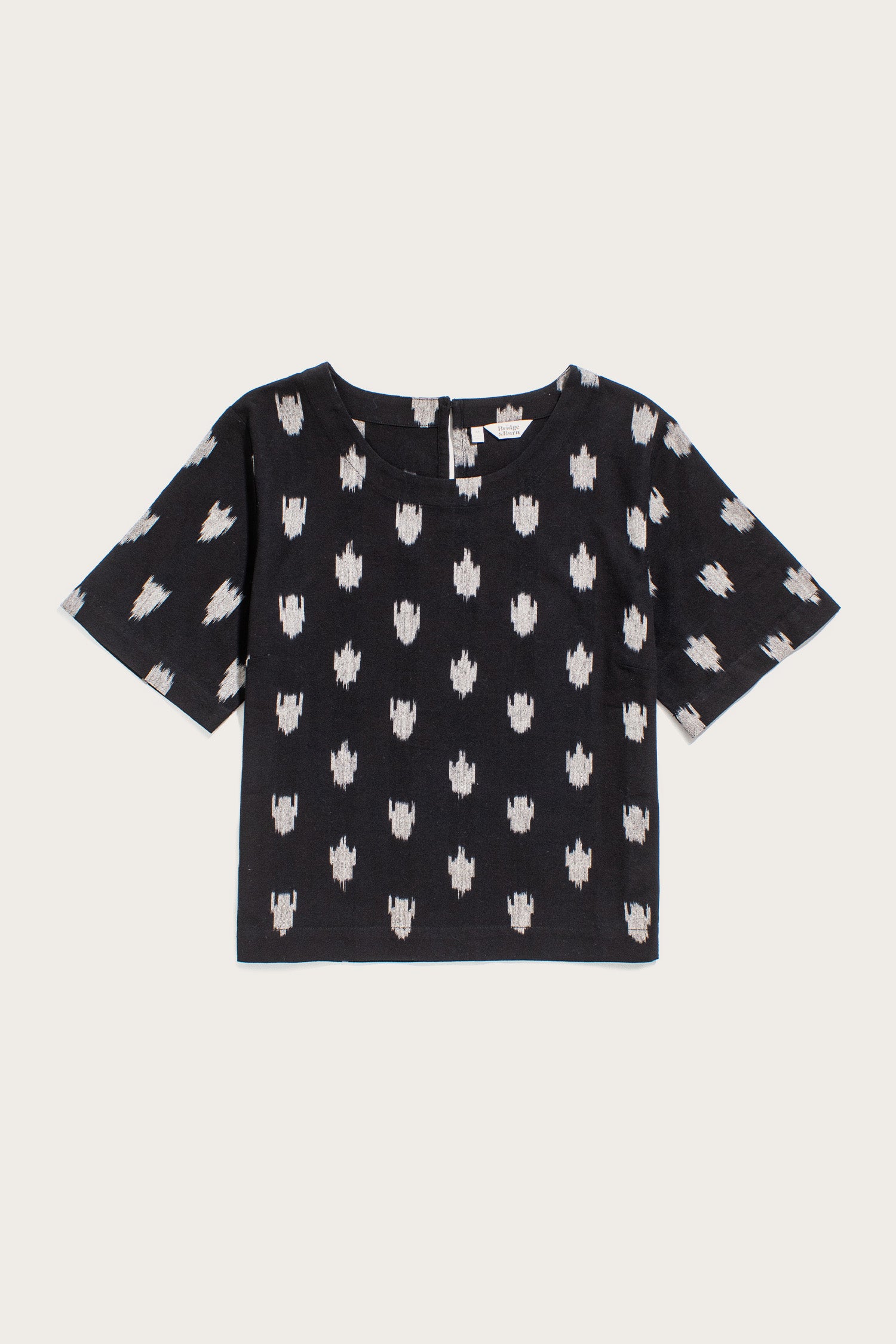 The Bridge & Burn Leona Shirt in Eclipse Ikat features short sleeves, a boxy fit, round neckline, and an evenly spaced white abstract pattern on black fabric. Displayed flat on a white background.