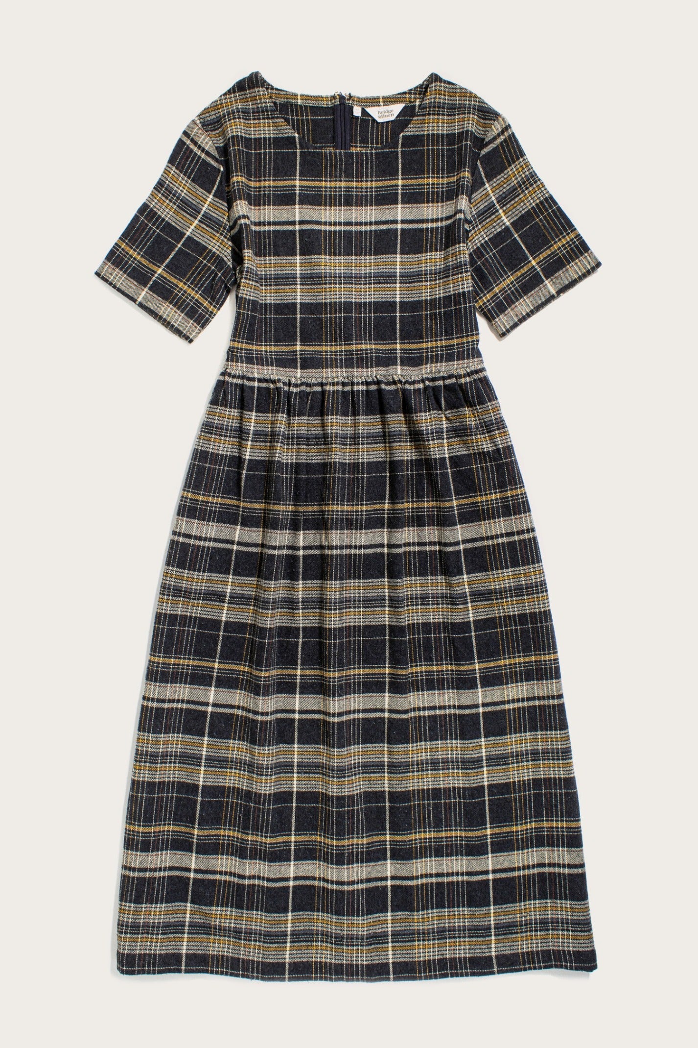 The Perry Dress in Volcanic Plaid by Bridge & Burn is a midi dress with short sleeves, made from recycled cotton. It features a dark navy, yellow, and white plaid pattern with a fitted bodice and gathered skirt.