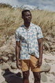 A man stands on a sandy beach with rocks and tall grass behind him, wearing the Bridge & Burn Bodie Shirt in Polar Blossom and tan shorts, gazing into the distance under a partly cloudy sky, effortlessly capturing summer style.