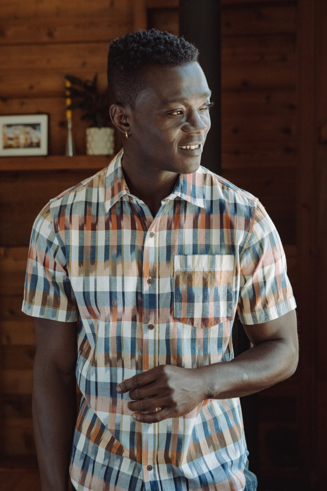A man indoors, smiling and looking to the side, wears the Bridge & Burn Harbor Slim Shirt in Aurora Space Dye Plaid. Wooden walls and a shelf with a framed photo and decor appear in the background.