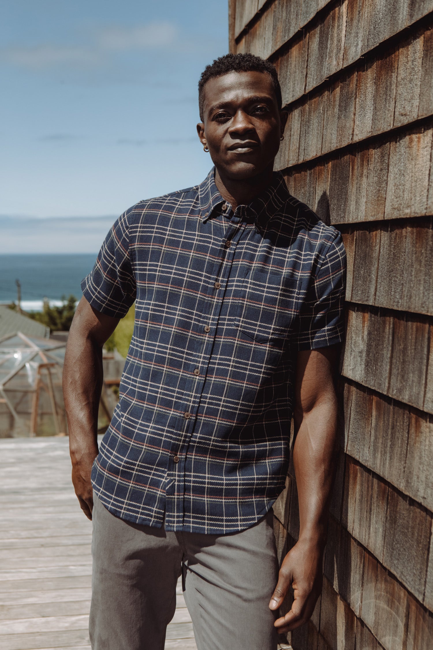 A man in the Bridge & Burn Jordan Slim Shirt in Dusk Herringbone Plaid and gray pants leans against a wooden shingle wall on a sunny deck, with glimpses of the ocean and a greenhouse in the background.