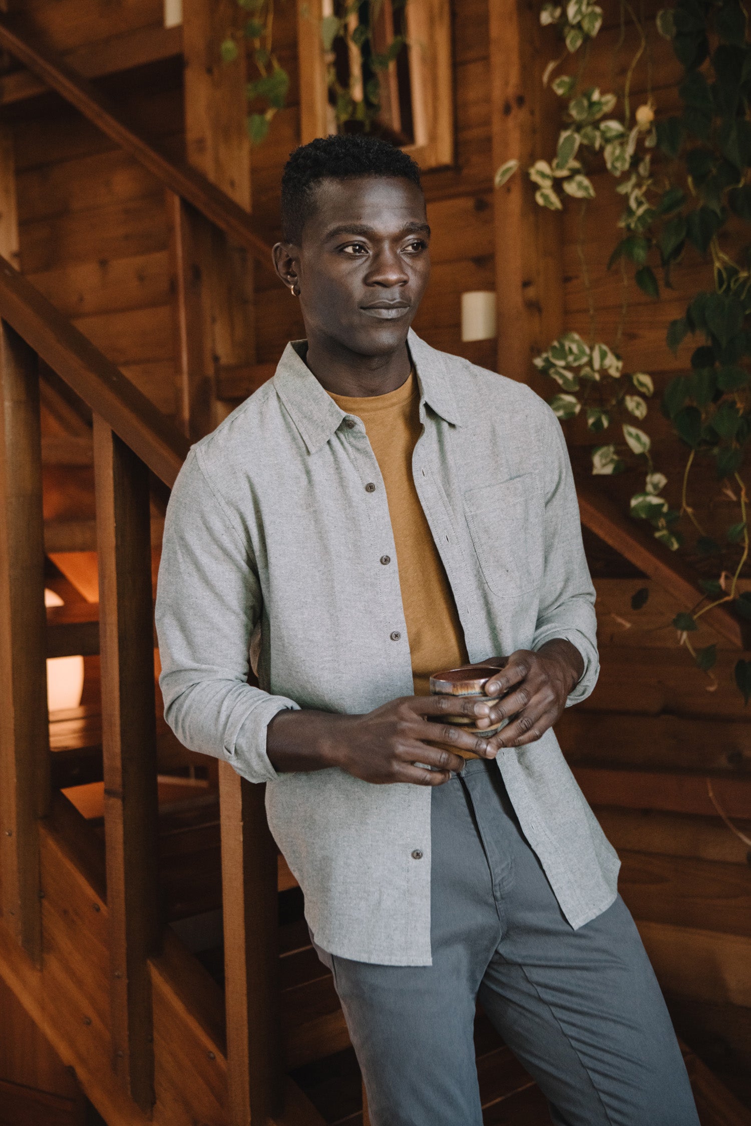 A man wearing the Bridge & Burn Larson Shirt in Sage Chambray stands by a wooden staircase and wall, holding a drink with green plants hanging nearby.