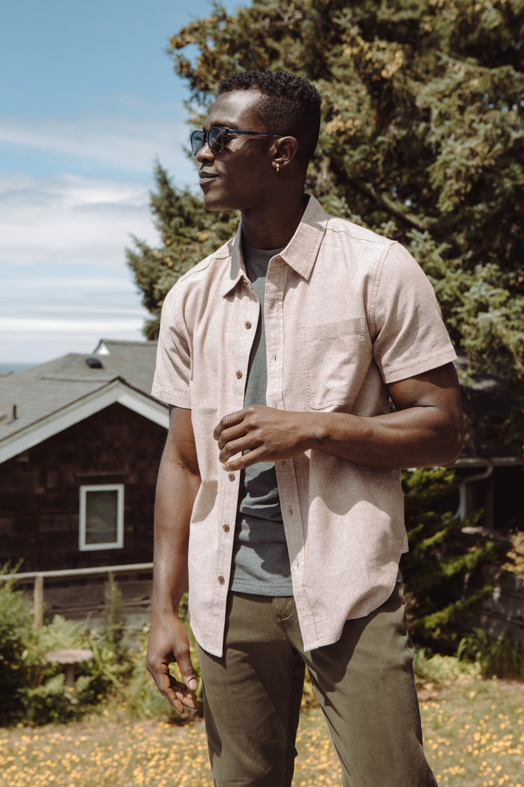 A man in sunglasses wears the Bridge & Burn Malcolm Shirt in Rust Chambray—an eco-friendly short sleeve button-up—over a gray tee with olive pants, standing outdoors on a sunny day with greenery and a house nearby.