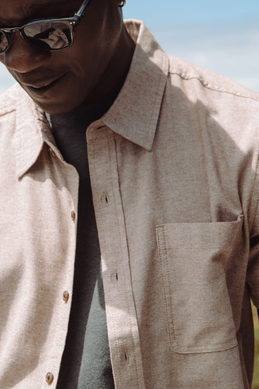 Someone wears the Bridge & Burn Malcolm Shirt in Rust Chambray outdoors, with sunglasses and only the lower face and upper body visible against a blurred background with blue sky.