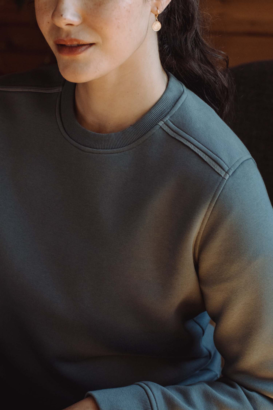 A woman with fair skin and brown hair in a ponytail wears a simple gold earring and the Bridge & Burn Bailey Crew Sweatshirt in Slate. Only her lower face and upper torso are visible as she sits indoors.
