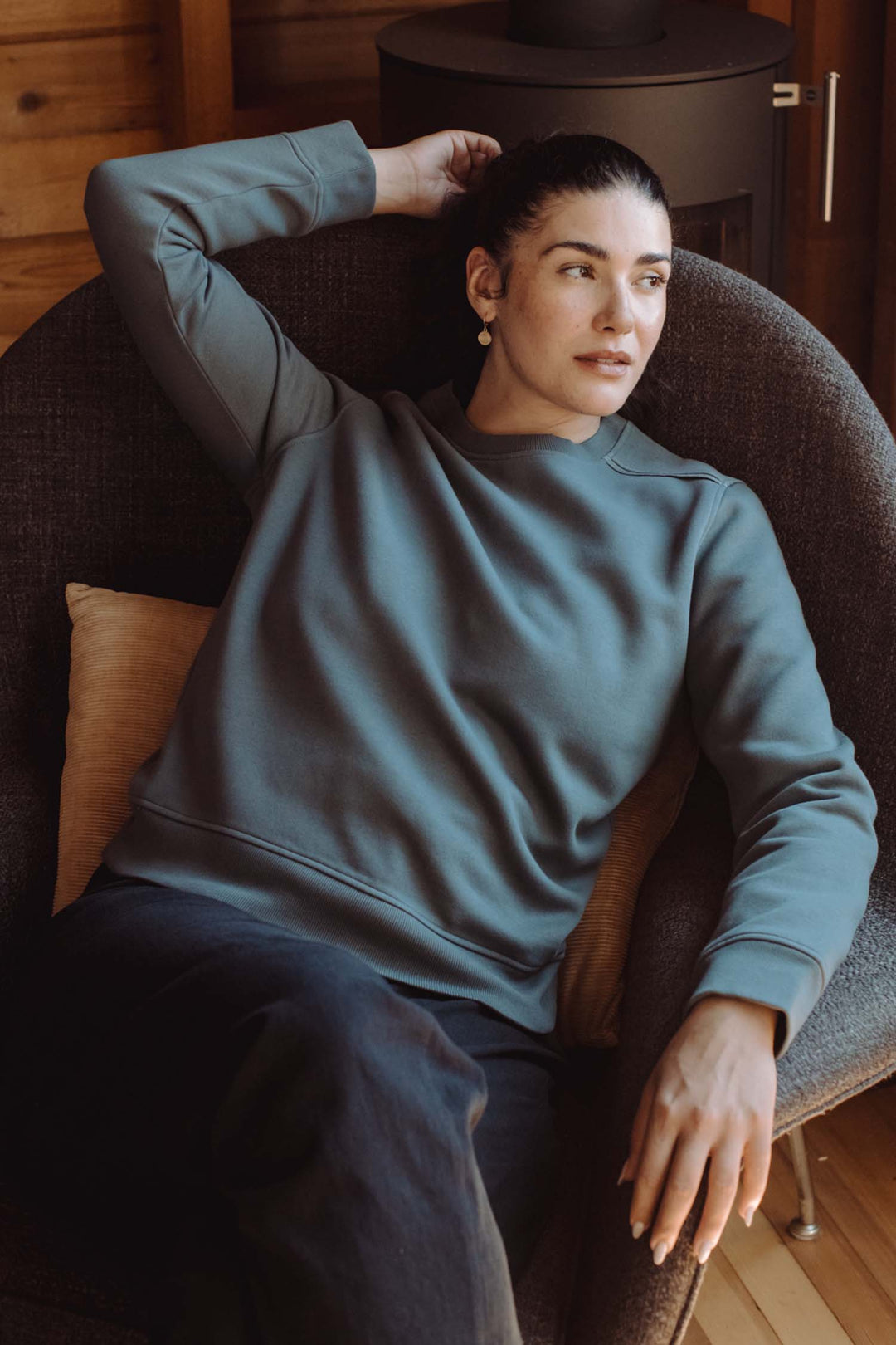A woman with dark hair tied back wears the Bridge & Burn Bailey Crew Sweatshirt in Slate and dark pants, relaxing indoors in a cushioned chair, resting one arm on the chair’s back and gazing thoughtfully to the side.